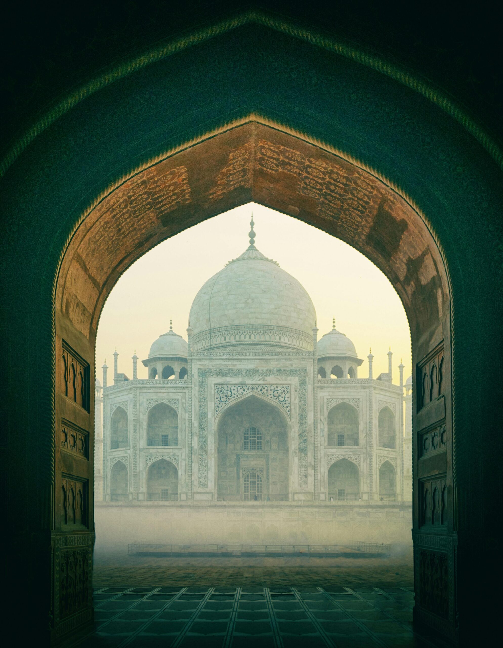 The iconic Taj Mahal viewed through a historic archway, enveloped in mist during sunrise in Agra, India.